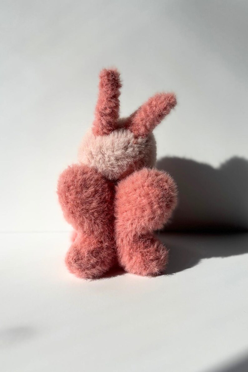May include: A plush bunny toy in shades of pink. The bunny has long, upright ears and a fluffy body. The fur is soft and textured, with a lighter pink on the face and a darker pink on the ears and body.