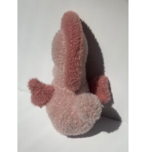 Op de afbeelding: Een roze pluche speelgoed met een lichtroze lichaam en donkerroze accenten op de oren en vleugels. Het speelgoed is gemaakt van zacht, pluizig materiaal en staat tegen een witte achtergrond.