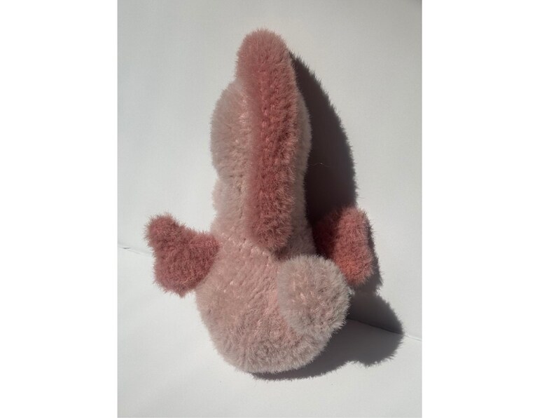 Op de afbeelding: Een roze pluche speelgoed met een lichtroze lichaam en donkerroze accenten op de oren en vleugels. Het speelgoed is gemaakt van zacht, pluizig materiaal en staat tegen een witte achtergrond.