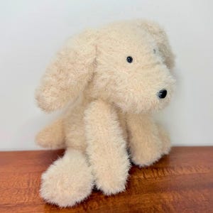 May include: A plush dog toy with shaggy, cream-colored fur. The stuffed animal features long, floppy ears, a small black nose, and two small black eyes. The toy is depicted in a seated position with its front paws resting forward.