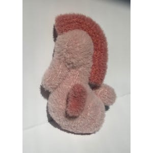 Op de afbeelding: Een pluche speelgoed in roze en roze tinten. Het speelgoed heeft een afgerond lichaam met een donkerroze accent op de rug en een roze mohawk-stijl detail bovenop. Het speelgoed is zacht en pluizig.