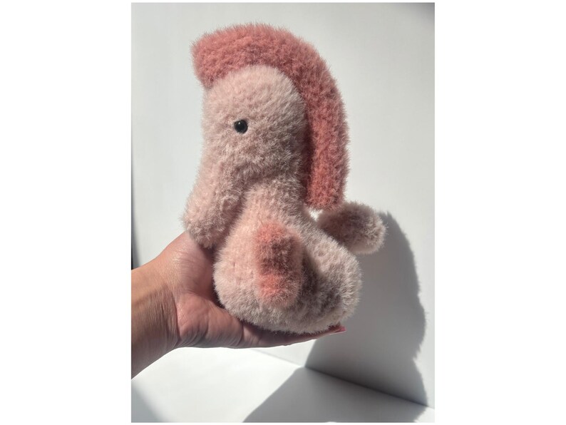 Op de afbeelding: Een handgemaakt pluche wezen in roze en roze tinten. Het zachte speelgoed heeft een getuften kuif en een afgerond lichaam. Het wezen wordt in een hand gehouden tegen een witte achtergrond, met een schaduw naar rechts.