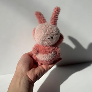 May include: A handmade plush bunny in shades of pink and white. The bunny has long, upright ears and a round body with striped detailing. The toy is held in a hand against a white background.
