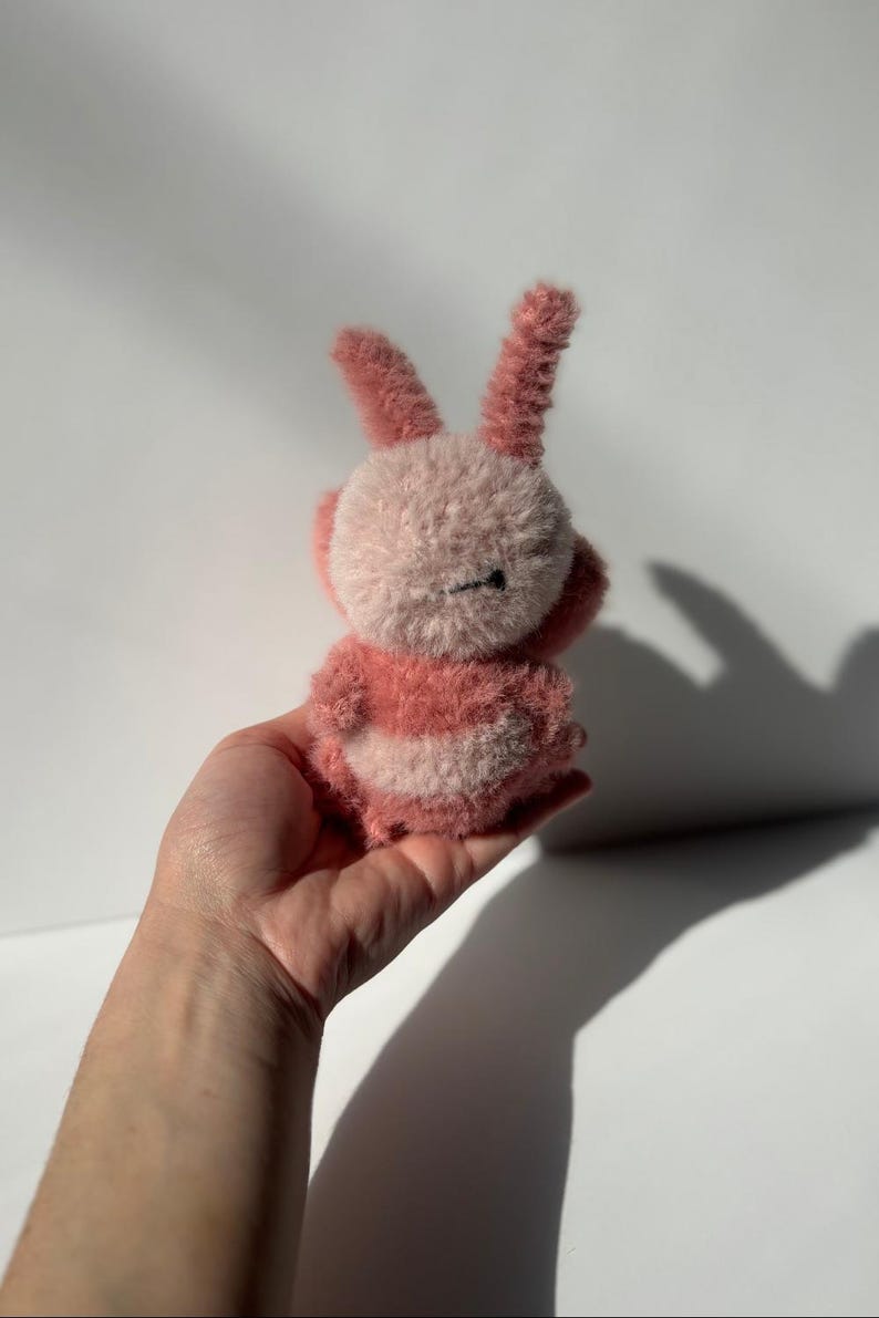 May include: A handmade plush bunny in shades of pink and white. The bunny has long, upright ears and a round body with striped detailing. The toy is held in a hand against a white background.