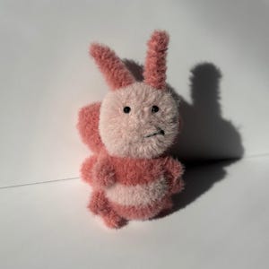 May include: A handmade plush bunny toy in shades of pink and white. The bunny has a fluffy body, long ears, and a slightly grumpy expression. The toy is made of soft, textured material, and is likely a children's toy or a decorative item.