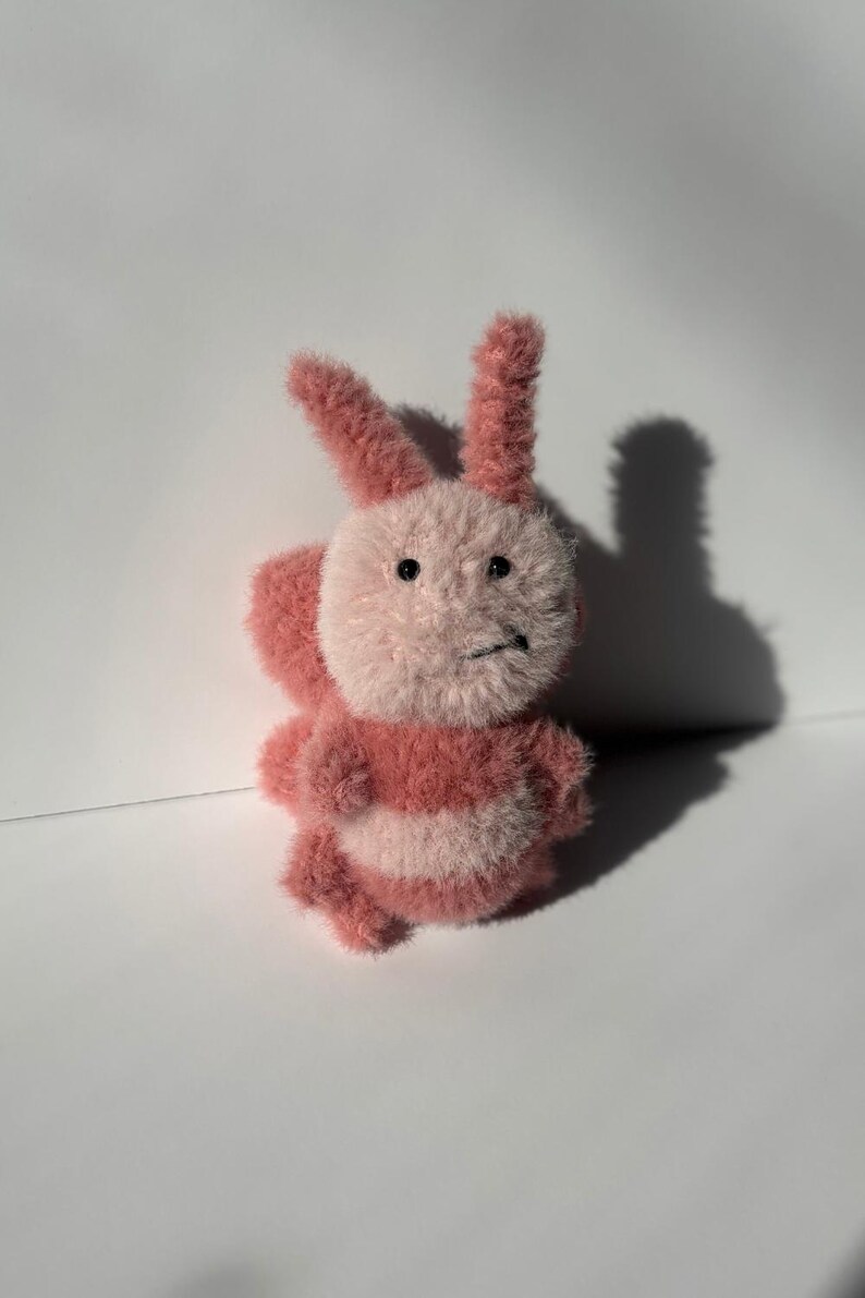 May include: A handmade plush bunny toy in shades of pink and white. The bunny has a fluffy body, long ears, and a slightly grumpy expression. The toy is made of soft, textured material, and is likely a children's toy or a decorative item.
