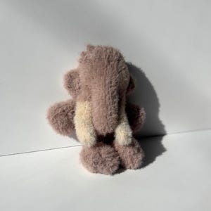 May include: A small, plush teddy bear in shades of mauve and cream, viewed from the back. The bear has a fluffy texture and a simple design, with visible arms, legs, and ears. The background is a plain white surface.