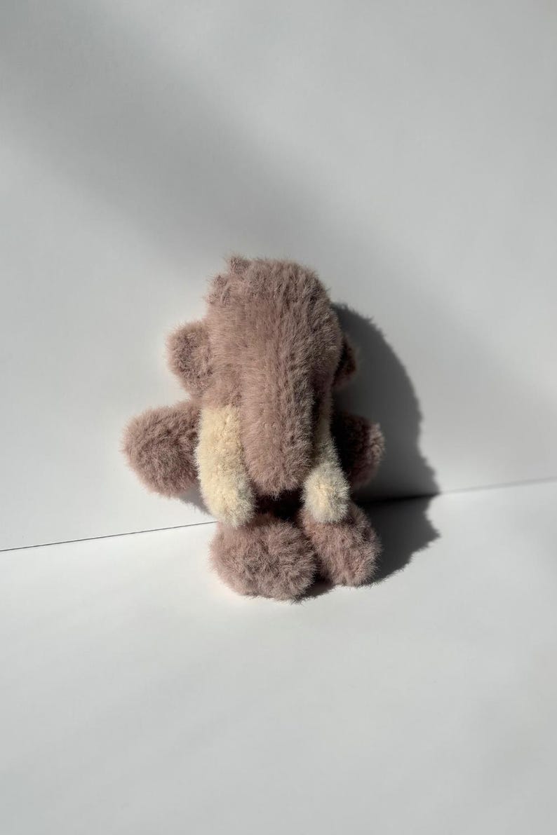 May include: A small, plush teddy bear in shades of mauve and cream, viewed from the back. The bear has a fluffy texture and a simple design, with visible arms, legs, and ears. The background is a plain white surface.