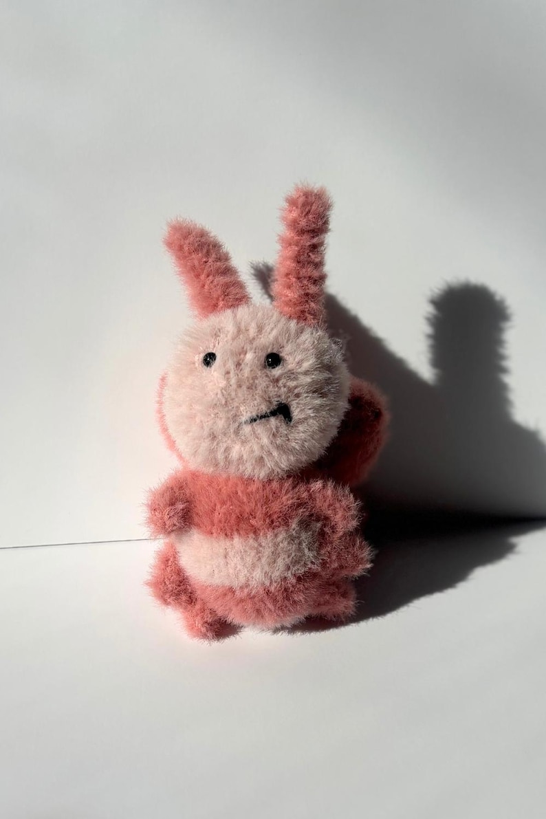 May include: A plush pink and white bunny toy with a fluffy texture. The bunny has upright ears, a round head, and a striped body. The toy is set against a white background, with a shadow cast to the right.