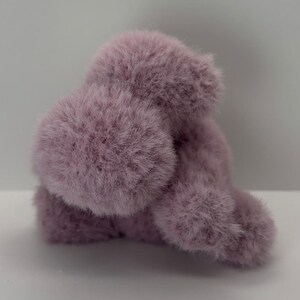 May include: A fluffy, lavender-colored plush toy, possibly a small animal or character. The toy has a soft, furry texture and a rounded shape, with visible tufts of fur. The background is a plain white surface.