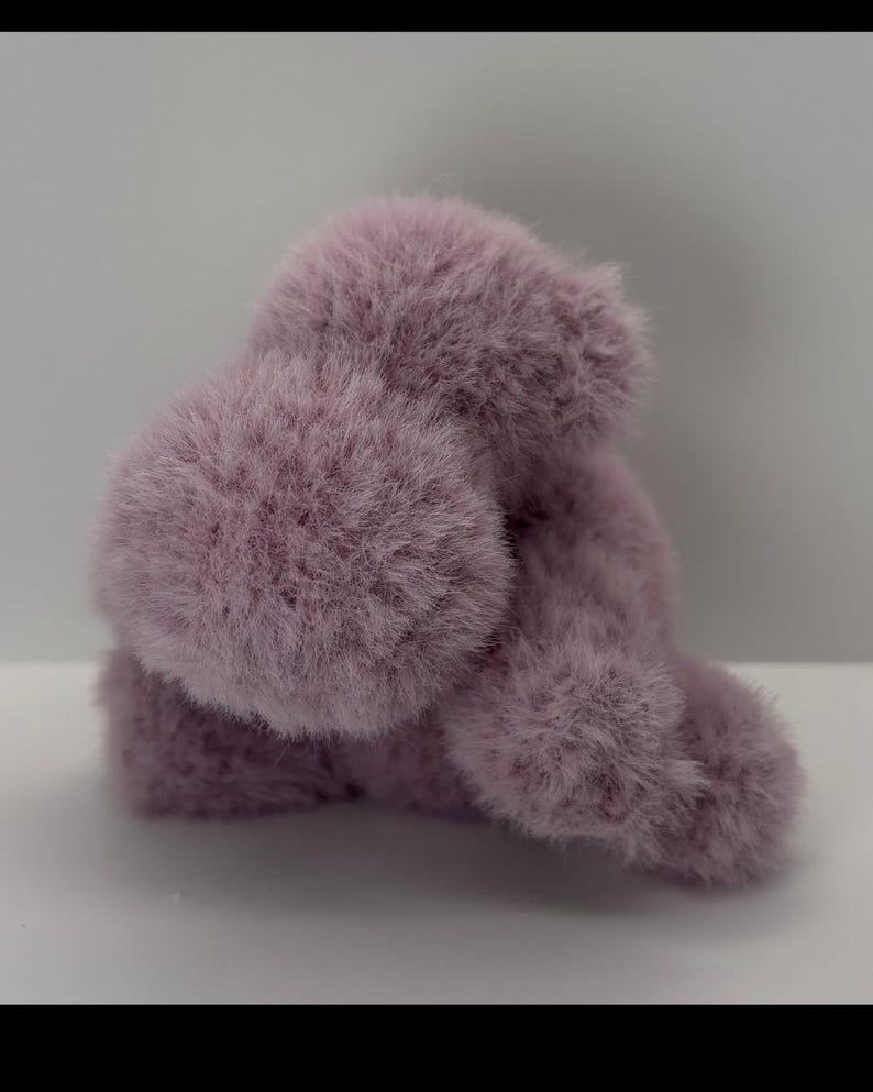 May include: A fluffy, lavender-colored plush toy, possibly a small animal or character. The toy has a soft, furry texture and a rounded shape, with visible tufts of fur. The background is a plain white surface.
