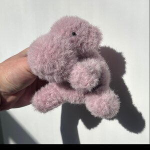 May include: A plush, lavender-colored stuffed animal with a fuzzy texture. The toy has a rounded body, small black eyes, and stubby limbs. The toy is designed to resemble a small, friendly creature.