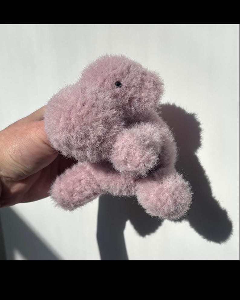 May include: A plush, lavender-colored stuffed animal with a fuzzy texture. The toy has a rounded body, small black eyes, and stubby limbs. The toy is designed to resemble a small, friendly creature.