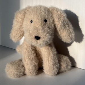 May include: A plush dog toy made of fuzzy, light beige yarn. The stuffed animal features long, floppy ears, a rounded snout, and two small, dark, glossy plastic eyes. A small, black, oval-shaped plastic nose is centered on the face of the toy.