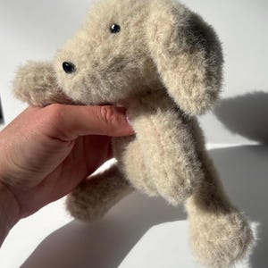 May include: A plush stuffed animal in the shape of a dog with light beige, fuzzy, textured fur. The toy features small, round, black plastic eyes and a black plastic nose. The dog has floppy ears and a soft, rounded body with four limbs.