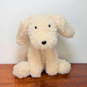 May include: A plush dog toy made of cream-colored, shaggy, textured fabric. The stuffed animal features long, floppy ears, a small black nose, and two dark, round eyes. It is depicted in a seated position with its front paws resting forward.