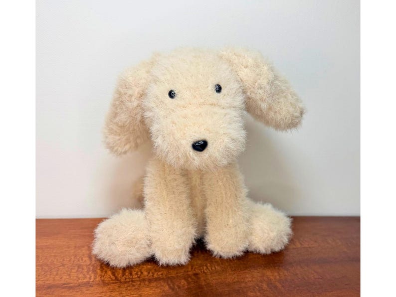 May include: A plush dog toy made of cream-colored, shaggy, textured fabric. The stuffed animal features long, floppy ears, a small black nose, and two dark, round eyes. It is depicted in a seated position with its front paws resting forward.