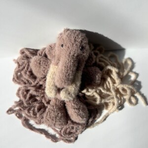 May include: A handmade plush toy in the shape of a crocodile, with a light brown body and a beige snout. The crocodile is surrounded by a pile of light brown yarn, creating a soft and textured appearance. The toy is set against a white background.