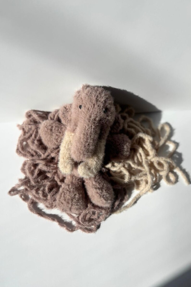 May include: A handmade plush toy in the shape of a crocodile, with a light brown body and a beige snout. The crocodile is surrounded by a pile of light brown yarn, creating a soft and textured appearance. The toy is set against a white background.