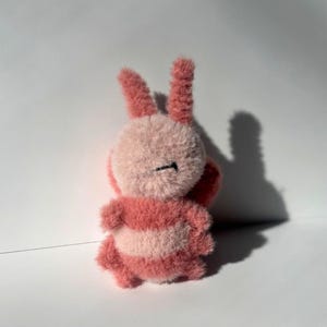 May include: A handmade plush bunny toy in shades of pink and white. The bunny has long, upright ears, a round body, and striped detailing. The toy is made of a soft, fuzzy material.