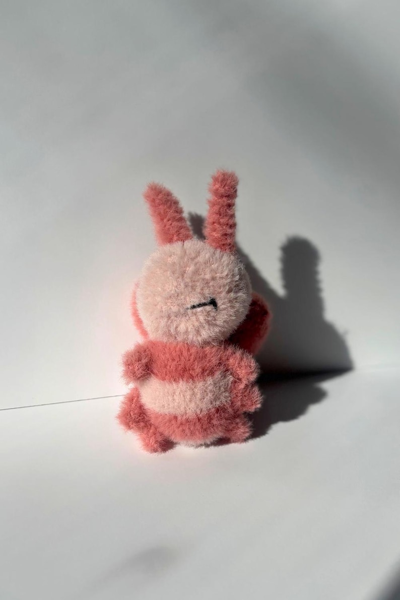May include: A handmade plush bunny toy in shades of pink and white. The bunny has long, upright ears, a round body, and striped detailing. The toy is made of a soft, fuzzy material.