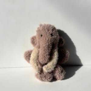 May include: A plush, lavender-colored elephant toy with a shaggy texture. The elephant has large, floppy ears and a trunk that curves downward. The tusks are a light beige color. The toy is sitting on a white surface.
