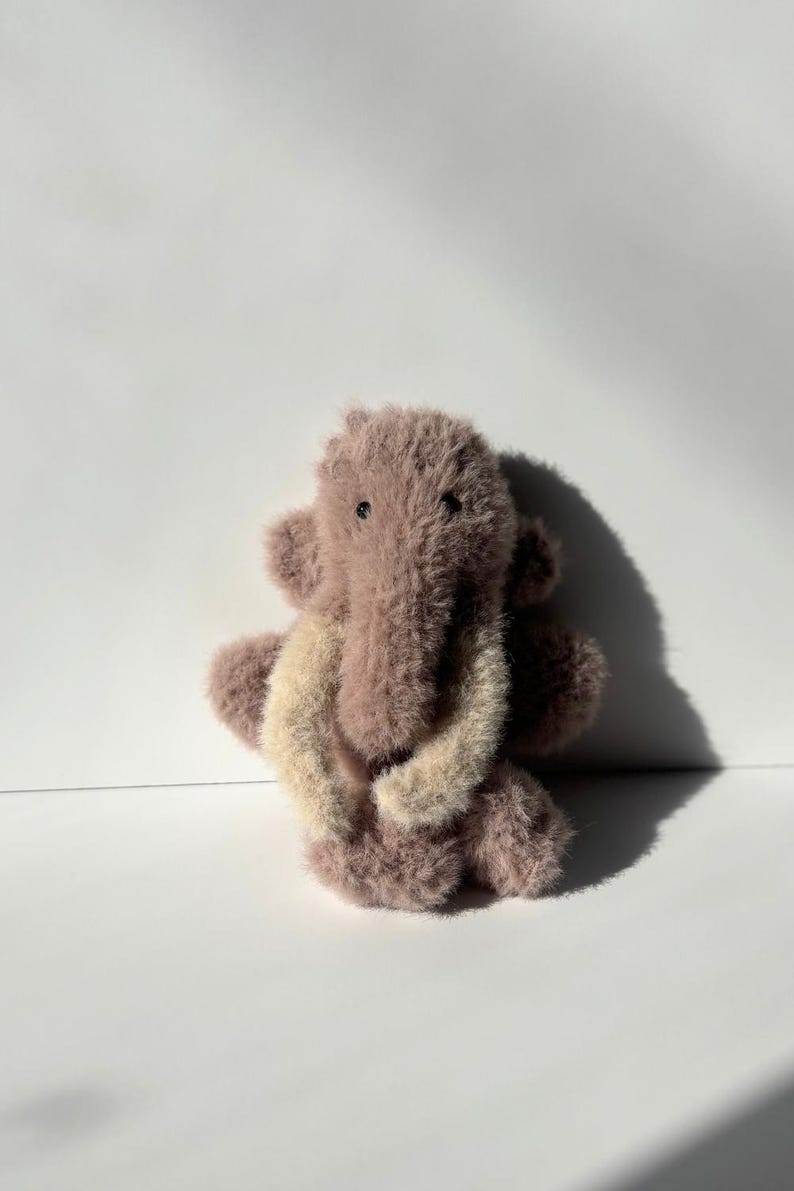 May include: A plush, lavender-colored elephant toy with a shaggy texture. The elephant has large, floppy ears and a trunk that curves downward. The tusks are a light beige color. The toy is sitting on a white surface.
