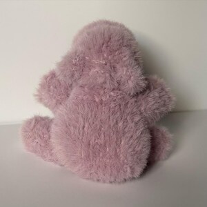 May include: A plush, lavender-colored stuffed animal with a fuzzy texture. The toy has a rounded body, small arms, and a head. The toy is sitting on a white surface.