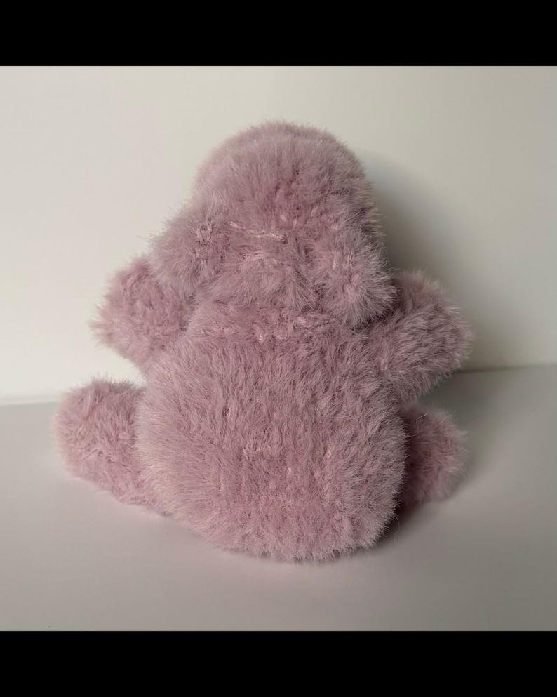 May include: A plush, lavender-colored stuffed animal with a fuzzy texture. The toy has a rounded body, small arms, and a head. The toy is sitting on a white surface.