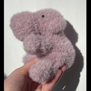 May include: A plush, lavender-colored stuffed animal shaped like a hippopotamus. The toy has a soft, fuzzy texture and a small, black eye. The hippo is held in a hand against a white background.