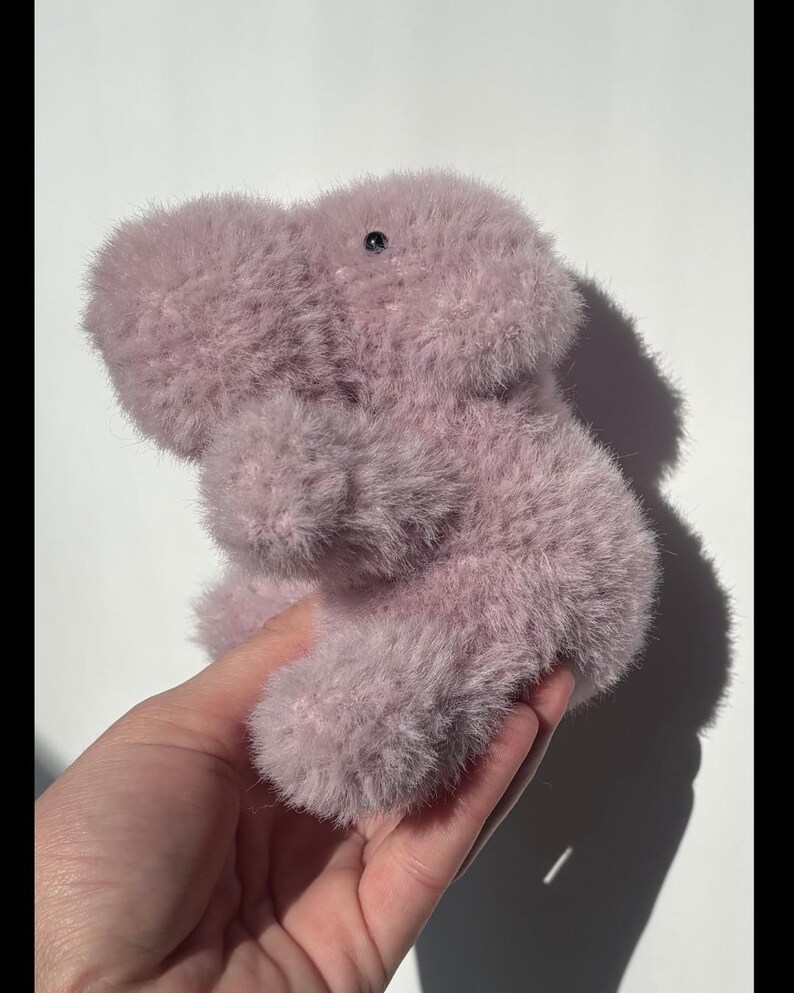 May include: A plush, lavender-colored stuffed animal shaped like a hippopotamus. The toy has a soft, fuzzy texture and a small, black eye. The hippo is held in a hand against a white background.