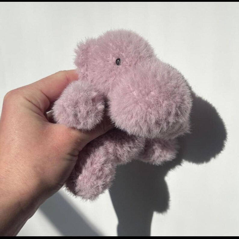 May include: A plush, lavender-colored hippo toy with a soft, furry texture. The toy has a small, black eye and is held against a white background. The hippo's shadow is visible.
