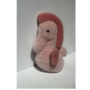 Op de afbeelding: Een pluche zeepaardje in roze en mauve tinten. Het zeepaardje heeft een getextureerd, pluizig lichaam en een donkerdere roze, rechtopstaande vin. Het heeft een klein, rond zwart oog en een gekrulde staart. Het speelgoed staat op een wit oppervlak.