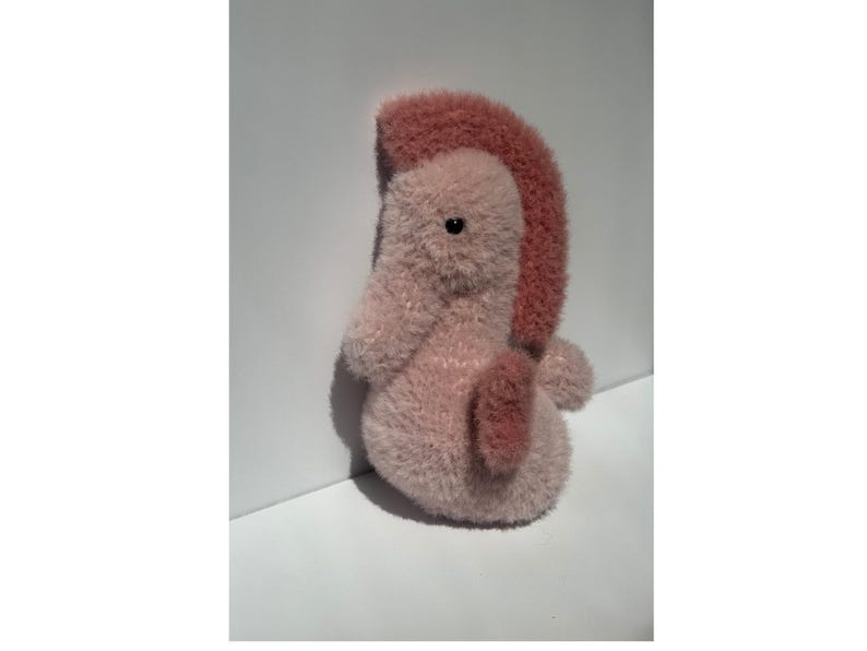 Op de afbeelding: Een pluche zeepaardje in roze en mauve tinten. Het zeepaardje heeft een getextureerd, pluizig lichaam en een donkerdere roze, rechtopstaande vin. Het heeft een klein, rond zwart oog en een gekrulde staart. Het speelgoed staat op een wit oppervlak.