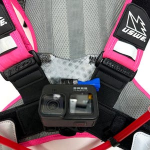 Op de afbeelding: Een roze en zwarte borsttuig met een GoPro-camera bevestigd aan de voorkant. Het harnas heeft een reflecterende strip op de schouderband en de tekst "USWE" is op de band gedrukt.