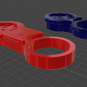 Puede incluir: Dos soportes de montaje para bicicleta impresos en 3D, uno rojo y otro azul. El soporte rojo tiene una abertura redonda y una superficie plana con un diseño en relieve. El soporte azul tiene dos aberturas redondas.