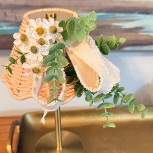 May include: A decorative table lamp with a woven lampshade, adorned with white daisy flowers and green foliage. The lamp has a gold-colored base and stem, and a cream-colored ribbon. The lamp is on a gold tray.