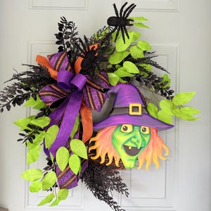 May include: A Halloween wreath featuring a witch's face with a purple hat and orange hair, surrounded by green leaves, black foliage, and a large purple and orange bow. Black spiders are also included in the design.