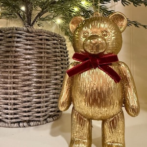 May include: A gold-colored teddy bear ornament with a textured surface and a dark red velvet ribbon tied around its neck. The bear has rounded ears, a small snout, and a gold string attached to the top of its head for hanging as a decoration.