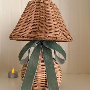 May include: A small, decorative lamp with a woven wicker shade and base. The shade is a light brown color, and the base is a rounded shape. A green velvet ribbon is tied around the base. A small, battery-operated candle is in the background.