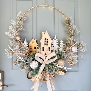 May include: A festive Christmas wreath with a gold metal hoop, adorned with faux pine branches, white and gold ornaments, and miniature wooden houses and trees. A large cream-colored bow adds a decorative touch. The wreath is lit with small, warm white lights.