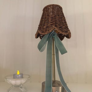 May include: A table lamp with a brown woven wicker lampshade featuring a scalloped edge and a sage green velvet ribbon tied in a bow. The lamp has a slender metallic gold stem and a circular base. Beside it sits a small clear glass bowl with a candle.