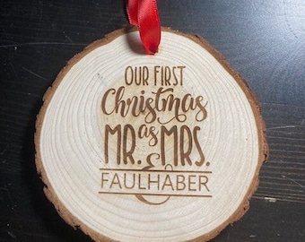 May include: A round wooden slice ornament with natural bark edges, featuring a red satin ribbon tied at the top. The surface is laser-engraved with the text: Our First Christmas as Mr. & Mrs. FAULHABER, centered in a mix of script and block fonts.