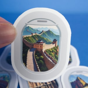May include: A white, oval-shaped sticker featuring an illustration of the Great Wall of China, viewed through a stylized airplane window. The artwork depicts the wall winding through a mountainous landscape with green trees and blue skies.