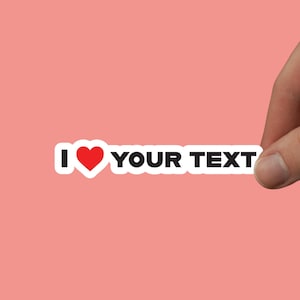 Op de afbeelding: Witte sticker met zwarte tekst die "Ik hou van JOUW TEKST" zegt met een rood hart in plaats van het woord "hou".