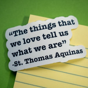 Puede incluir: Una pegatina blanca con borde festoneado, con la cita "Las cosas que amamos nos dicen lo que somos" de Santo Tomás de Aquino en texto negro. La pegatina está sobre un papel rayado amarillo, con un fondo verde.