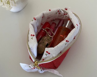 May include: A red and white cherry print makeup bag with a gold zipper. The bag is open and filled with various beauty products, including a red lipstick, a clear bottle of hair oil, and a silver keychain.