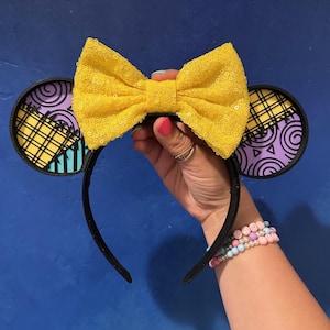 May include: Black headband with mouse ears featuring a yellow sequined bow. The ears have a circular design with purple, yellow, and black patterns. A fun accessory for themed events.