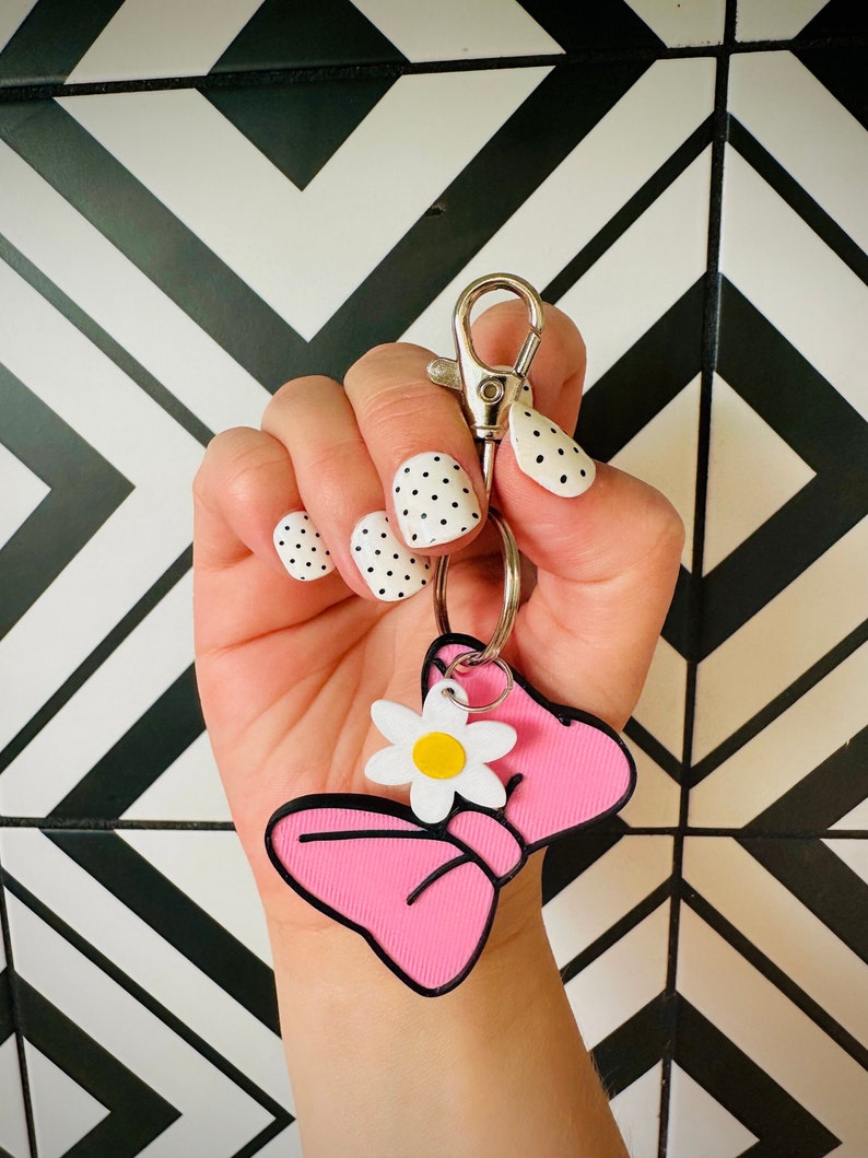 May include: A pink bow keychain with a white daisy charm. The keychain is attached to a silver key ring. The keychain is held in a hand with white nail polish with black polka dots.