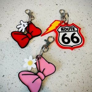 May include: Three keychains with different designs. One is a red bow with a white Mickey Mouse hand, another is a pink bow with a white daisy, and the last is a red Route 66 sign with a yellow lightning bolt.